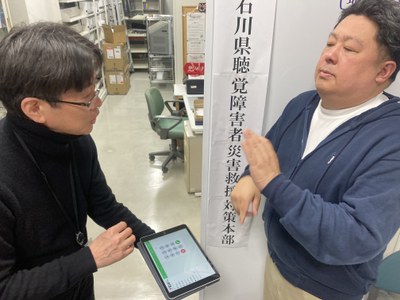 日本財団電話リレーサービススタッフが石川県聴覚障害者協会スタッフへ電話リレーサービスの入ったiPadを渡している写真